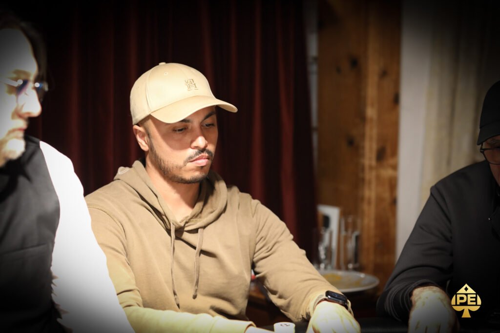 Ein Mann sitzt an einem Pokertisch und hält seine Karten in der Hand. Er hat einen konzentrierten Gesichtsausdruck und trägt eine schwarze Kappe sowie ein dunkles Hemd. Im Hinter