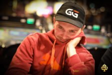 Ein Mann sitzt an einem Pokertisch, umgeben von anderen Spielern. Er hält Karten in der Hand und schaut konzentriert auf den Tisch. Die Atmosphäre ist gespannt und fokussiert
