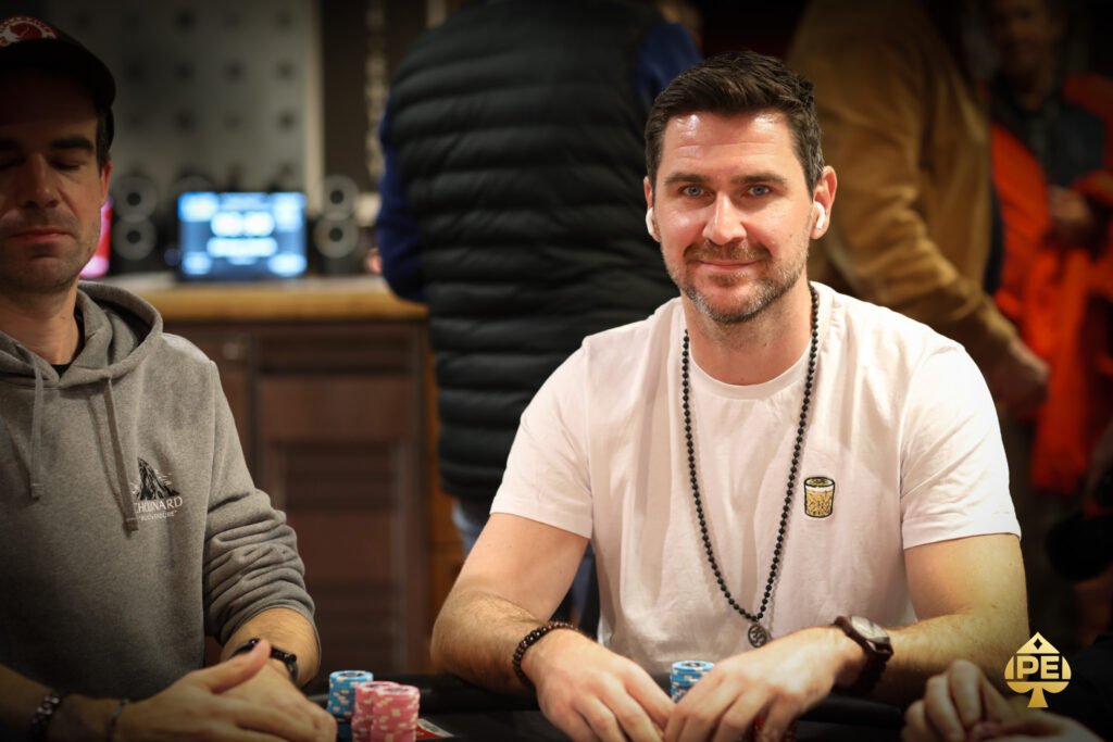 Ein Mann in einem dunklen Anzug und einer Fliege sitzt an einem Pokertisch und hält zwei Karten in der Hand. Er schaut konzentriert auf den Tisch, wo Pokerchips und Karten lie