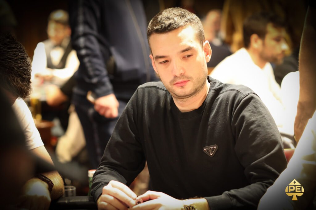 Ein Mann sitzt an einem Pokertisch, umgeben von anderen Spielern. Er hält Karten in der Hand und schaut konzentriert auf den Tisch. Die Szene spielt sich in einem eleganten Pokerraum