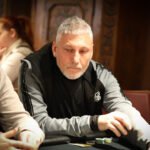 Ein professioneller Pokerspieler mit Sonnenbrille und Kappe sitzt an einem Pokertisch und betrachtet konzentriert seine Karten. Im Hintergrund sind andere Spieler und Chips zu sehen.
