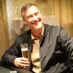 Ein Mann in einem schwarzen Anzug und einer Sonnenbrille sitzt an einem Pokertisch und hält seine Karten fest in der Hand, während er ernst und konzentriert wirkt. Andere Spieler und Chips sind