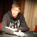 Ein Mann und eine Frau sitzen an einem Pokertisch und spielen Karten. Der Mann hat einen ernsten Blick und trägt eine dunkle Brille, während die Frau lächelt und ihre Karten betrachtet. Im Hinter