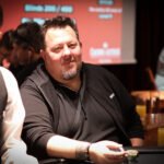 Ein Mann in einem schwarzen Anzug und einer schwarzen Sonnenbrille sitzt vor einem Pokertisch und hält Chips in der Hand. Im Hintergrund sind andere Pokerspieler und Spielkarten zu sehen.