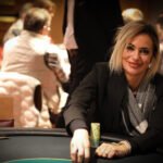 Ein Mann in einem Anzug sitzt an einem Pokertisch und hält seine Spielkarten in der Hand. Auf dem Tisch liegen Pokerchips und eine Glaskaraffe mit Wasser. Im Hintergrund sind weitere Spieler und