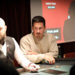 Ein Mann mit Bart und Brille sitzt an einem Pokertisch und hält Karten in der Hand. Er wirkt konzentriert und schaut ernst in die Kamera. Hinter ihm ist eine verschwommene Gl