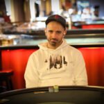Ein Mann sitzt an einem Pokertisch und hält seine Karten in der Hand, während er konzentriert auf seine Gegner blickt. Die Chips vor ihm stapeln sich hoch.
