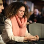 Ein Mann sitzt an einem Pokertisch und hält zwei Spielkarten in der Hand. Er trägt einen dunklen Anzug und eine schwarze Krawatte. Im Hintergrund sind Pokerchips und andere Spieler zu sehen.