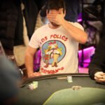 Ein Mann in einem dunkelblauen Anzug und einer Krawatte steht vor einem Pokertisch. Er hält eine Hand mit Spielkarten und schaut konzentriert auf die Karten. Im Hintergrund sind weitere Spieler