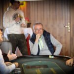 Ein grüner Pokertisch mit Spielkarten, Pokerchips und Getränken darauf, umgeben von Spielern in einer entspannten Atmosphäre.