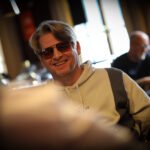 Ein Mann und eine Frau sitzen an einem Pokertisch und spielen eine Partie Poker. Der Mann hat eine Hand mit Spielkarten in der Luft und schaut konzentriert auf den Tisch, während die Frau Karten auf der