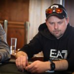 Ein Mann in einem schwarzen Anzug und einer roten Krawatte sitzt an einem Pokertisch und hält seine Spielkarten in der Hand. Sein Gesichtsausdruck ist konzentriert, während er sein