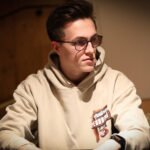 Ein Mann in einem eleganten Anzug sitzt an einem Pokertisch und hält seine Spielkarten in der Hand, während er konzentriert auf seine Gegner schaut. Die Atmosphäre ist gespannt und die Be