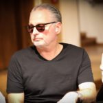 Ein Mann sitzt an einem Pokertisch und hält zwei Spielkarten in der Hand, während er konzentriert auf seine Gegner schaut. Die Karten und Chips auf dem Tisch deuten auf ein intensives Pokers