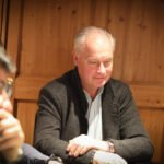 Ein Mann in einem eleganten Anzug und einer schwarzen Sonnenbrille sitzt an einem Pokertisch und hält seine Spielkarten in der Hand. Im Hintergrund sind weitere Spieler und Pokerchips zu sehen.