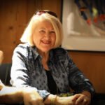Ein Mann und eine Frau sitzen eng beieinander an einem Pokertisch. Der Mann hält Pokerchips in der Hand, während die Frau konzentriert auf ihre Karten schaut. Im Hintergrund sind weitere Spieler