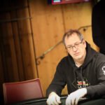 Ein Mann sitzt an einem Pokertisch mit grünem Filz und hält Karten in der Hand. Er hat einen nachdenklichen Ausdruck und trägt einen dunklen Anzug. Im Hintergrund sind Pokerch