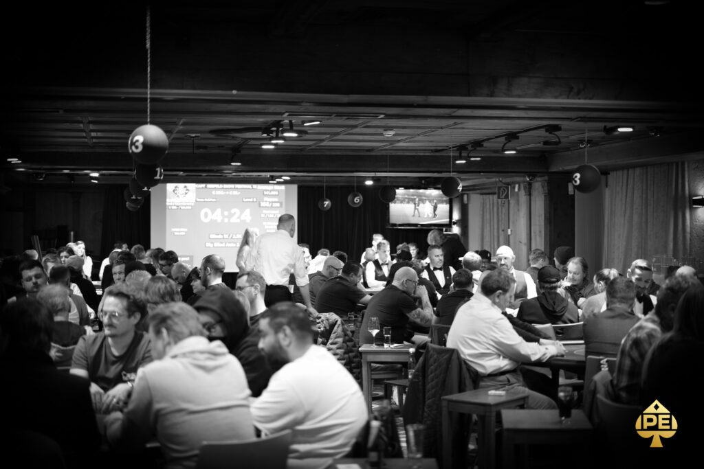 Ein Mann in einem schwarzen Anzug sitzt an einem Pokertisch, umgeben von Spielkarten, Pokerchips und einem Stapel Jetons. Er hält eine Hand mit Karten und hat einen konzentrierten Blick auf
