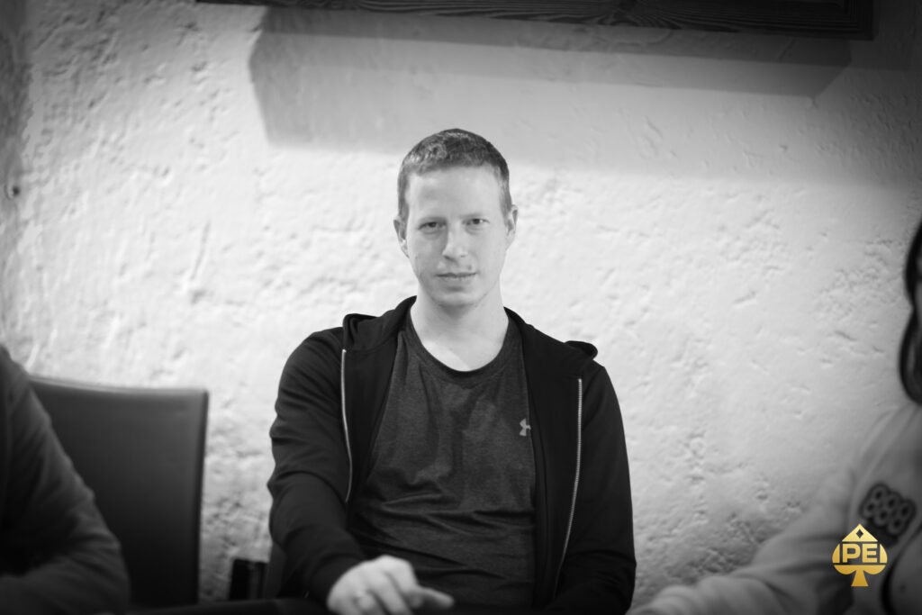 Ein Mann in einem eleganten Anzug sitzt an einem Pokertisch und hält seine Karten in der Hand, während er konzentriert auf die Mitte des Tisches schaut. Im Hintergrund sind andere Spieler und Poker