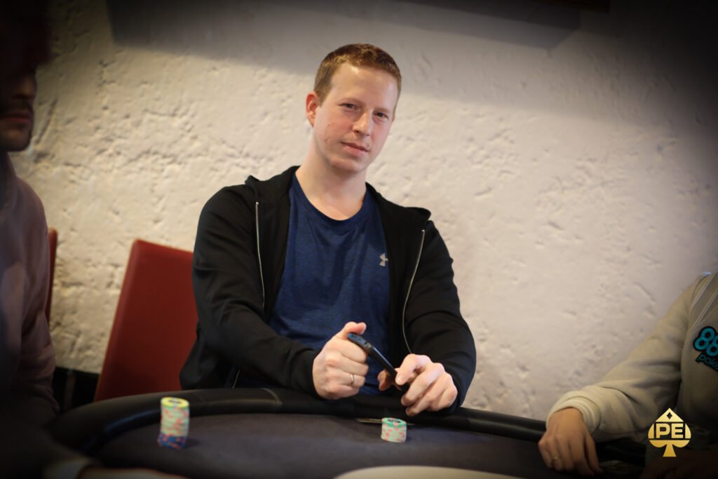 Ein Mann in einem dunkelblauen Anzug und einer roten Krawatte sitzt an einem Pokertisch und hält seine Karten in der Hand. Er hat einen konzentrierten Blick und ein selbstbewusstes L
