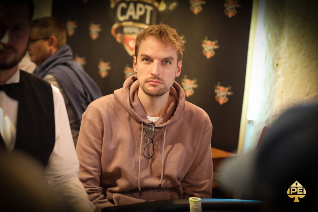 Ein Mann in einem dunkelblauen Anzug und einer roten Krawatte sitzt an einem Pokertisch und hält zwei Spielkarten in der Hand. Er schaut konzentriert auf den Tisch, umgeben von