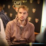 Ein Mann in einem dunkelblauen Anzug und einer roten Krawatte sitzt an einem Pokertisch und hält zwei Spielkarten in der Hand. Er schaut konzentriert auf den Tisch, umgeben von