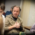 Ein Mann und eine Frau sitzen an einem Pokertisch und spielen Poker. Der Mann hält Karten in der Hand und schaut konzentriert auf den Tisch, während die Frau ihm gegenüber mit ernstem Blick ihre Chips betr