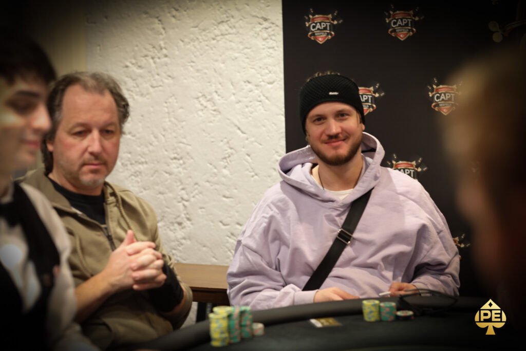 Ein Mann in einem schwarzen Anzug und einer schwarzen Sonnenbrille sitzt an einem Pokertisch und hält Karten in der Hand, während er konzentriert auf seine Gegner schaut.