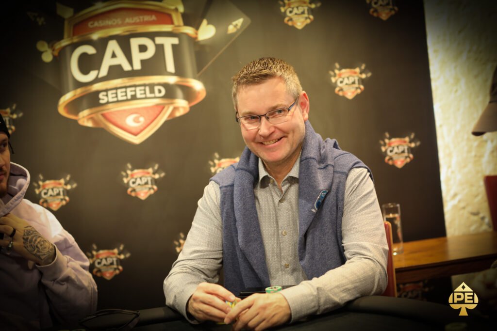 Ein Mann sitzt an einem Pokertisch und hält zwei Spielkarten in der Hand. Er wirkt konzentriert und nachdenklich, während er auf die Karten schaut. Im Hintergrund sind Pokerchips