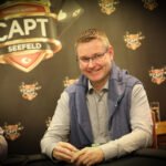 Ein Mann sitzt an einem Pokertisch und hält zwei Spielkarten in der Hand. Er wirkt konzentriert und nachdenklich, während er auf die Karten schaut. Im Hintergrund sind Pokerchips