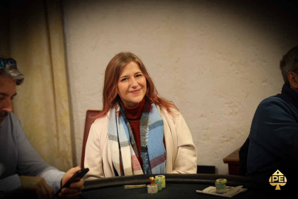 Ein Mann in einem dunkelblauen Anzug sitzt an einem Pokertisch mit grünem Filz. Er hält Karten in der Hand und schaut konzentriert auf den Tisch. Im Hintergrund sind Poker