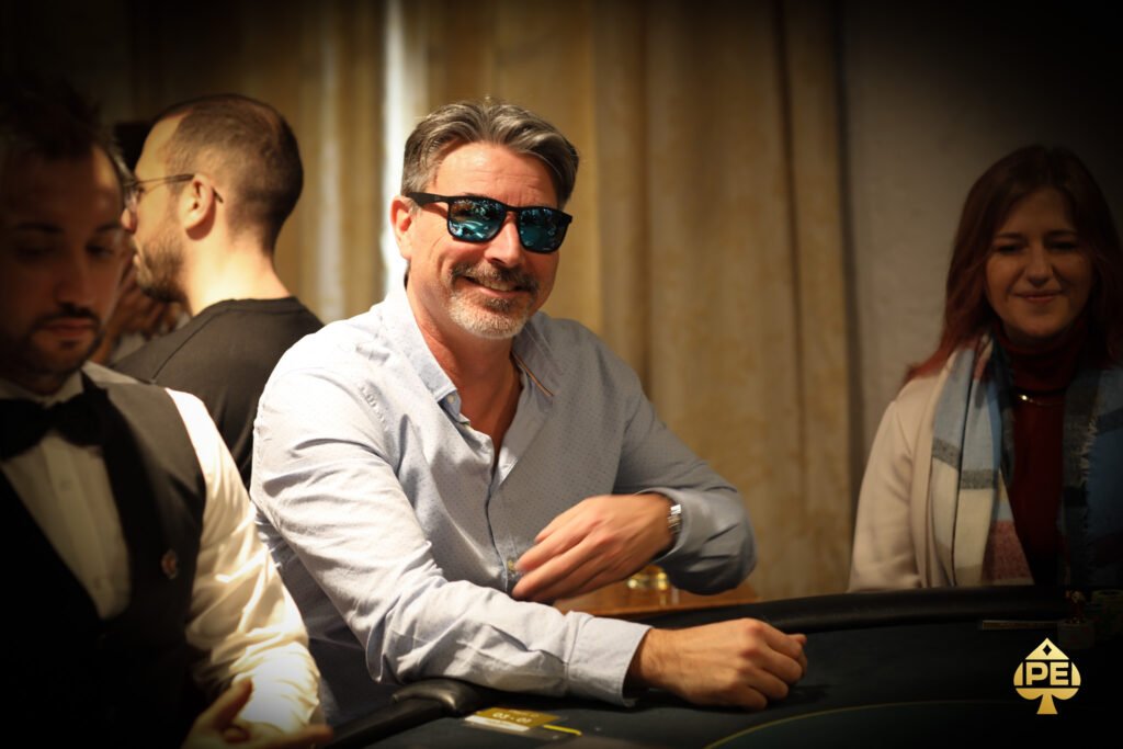 Ein Mann in einem Anzug spielt Poker an einem eleganten Tisch in einem stilvollen Casino. Er hält Karten in der Hand und schaut konzentriert auf den Tisch. Andere Spieler sind im Hintergrund