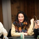 Ein Mann sitzt an einem Pokertisch und hält zwei Spielkarten in der Hand. Er trägt einen dunklen Anzug und hat eine ernste Miene. Um ihn herum sind Pokerchips und Gläser zu sehen