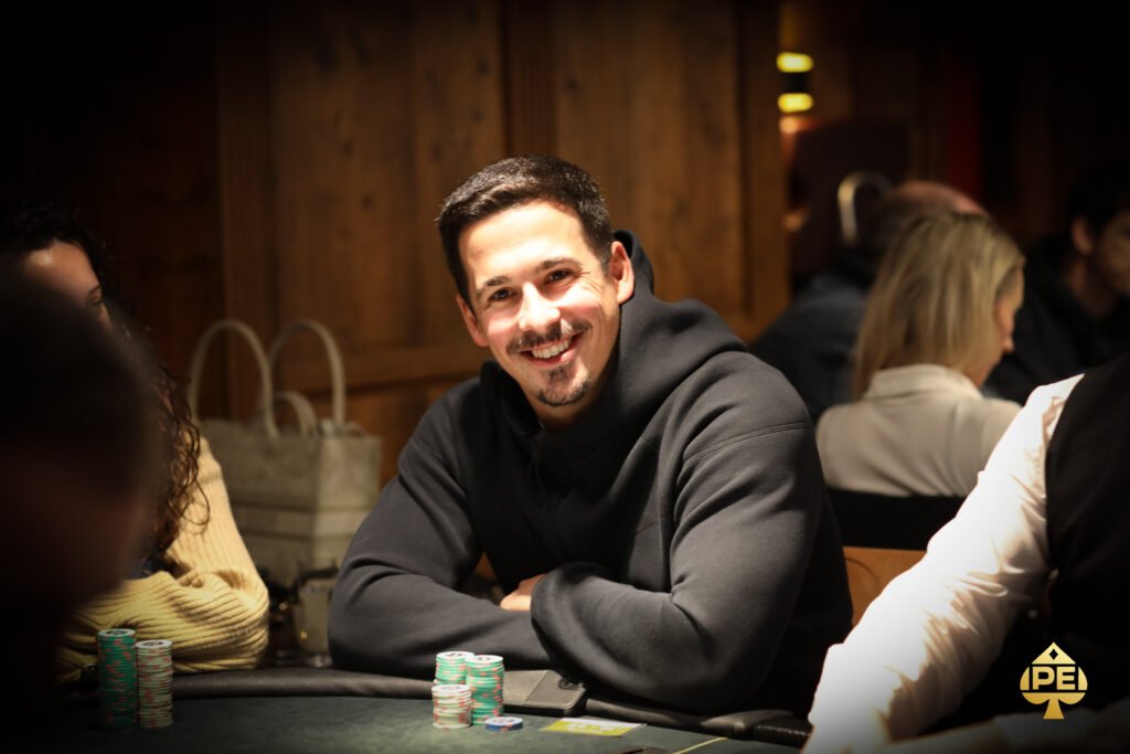Ein Mann sitzt an einem Pokertisch mit grünem Filz und hält Karten in der Hand. Im Hintergrund sind Chips und andere Spieler zu sehen.