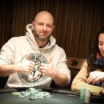 Ein Mann sitzt an einem Pokertisch und betrachtet seine Karten mit ernstem Gesichtsausdruck, umgeben von anderen Spielern. Die Atmosphäre ist konzentriert und spannungsgeladen.