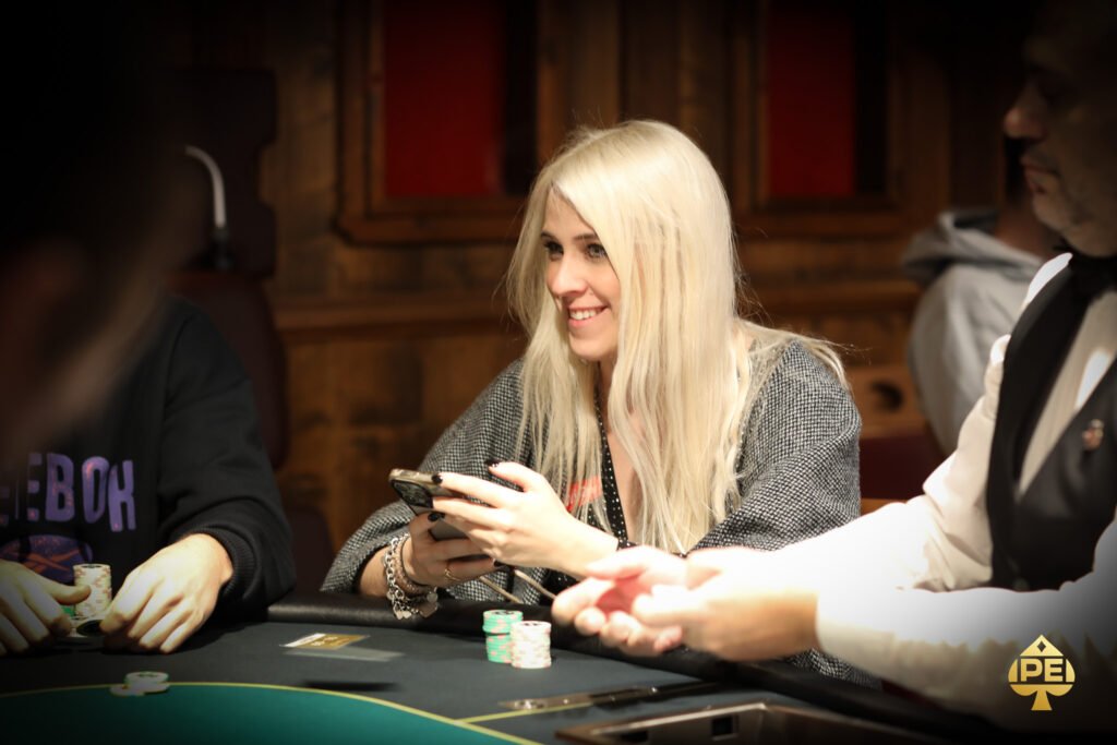 Ein Mann sitzt an einem Pokertisch und hält zwei Spielkarten in der Hand. Er hat eine ernste Miene und trägt einen dunklen Anzug. Im Hintergrund sind Casino-Chips und weitere Spieler zu sehen.