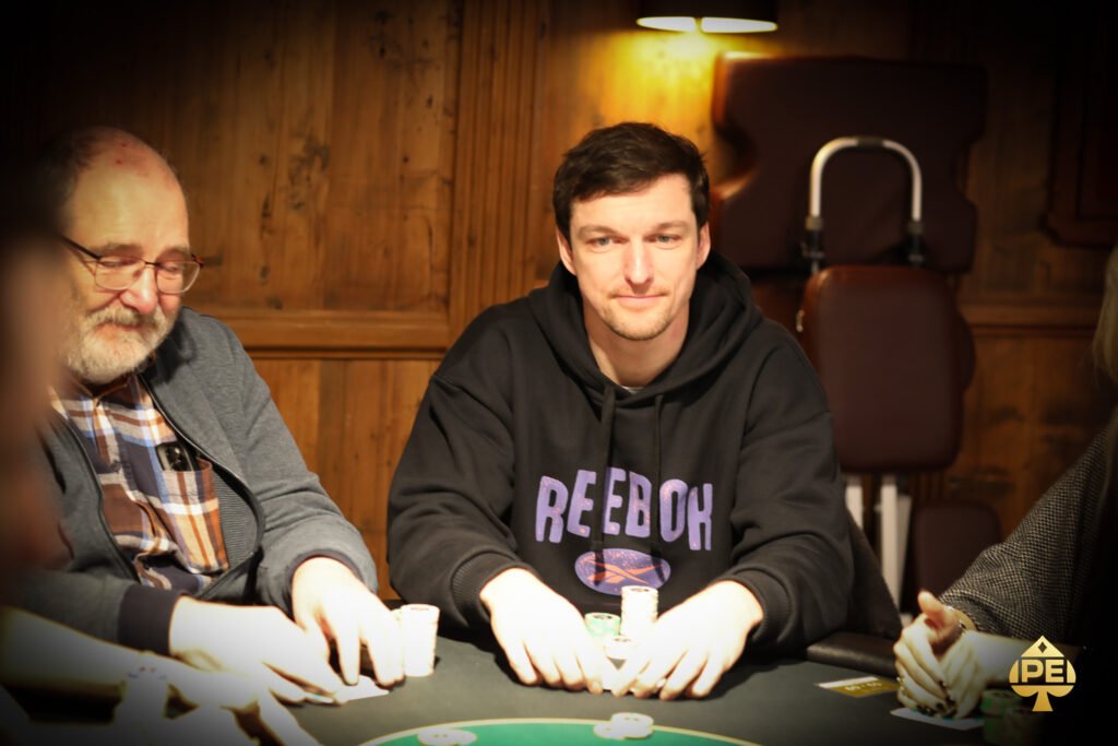 Ein Mann sitzt an einem Pokertisch und hält Karten in der Hand, während er konzentriert auf seine Gegner blickt. Im Hintergrund sind Pokerchips und Getränke zu sehen.
