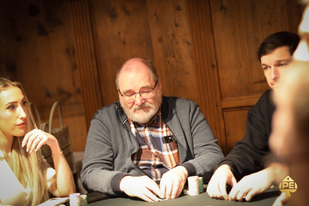 Ein Mann sitzt an einem Pokertisch und hält Karten in der Hand. Er hat eine konzentrierte Miene und trägt einen Anzug. Im Hintergrund sind weitere Spieler zu sehen.