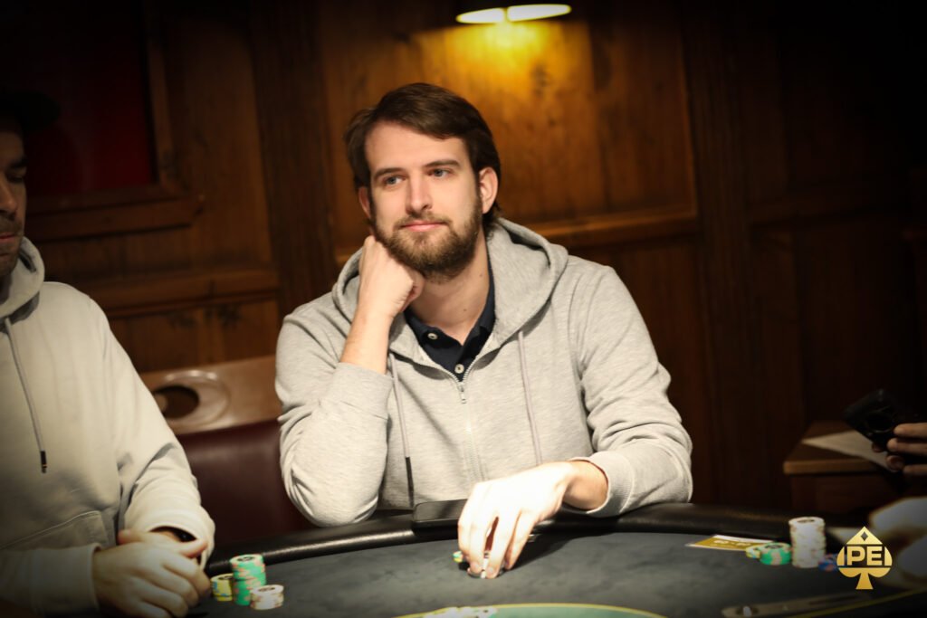 Ein Mann in einem schwarzen Anzug und einer schwarzen Sonnenbrille spielt Poker an einem Tisch, umgeben von anderen Spielern. Er hält Karten in der Hand und schaut konzentriert auf den Tisch