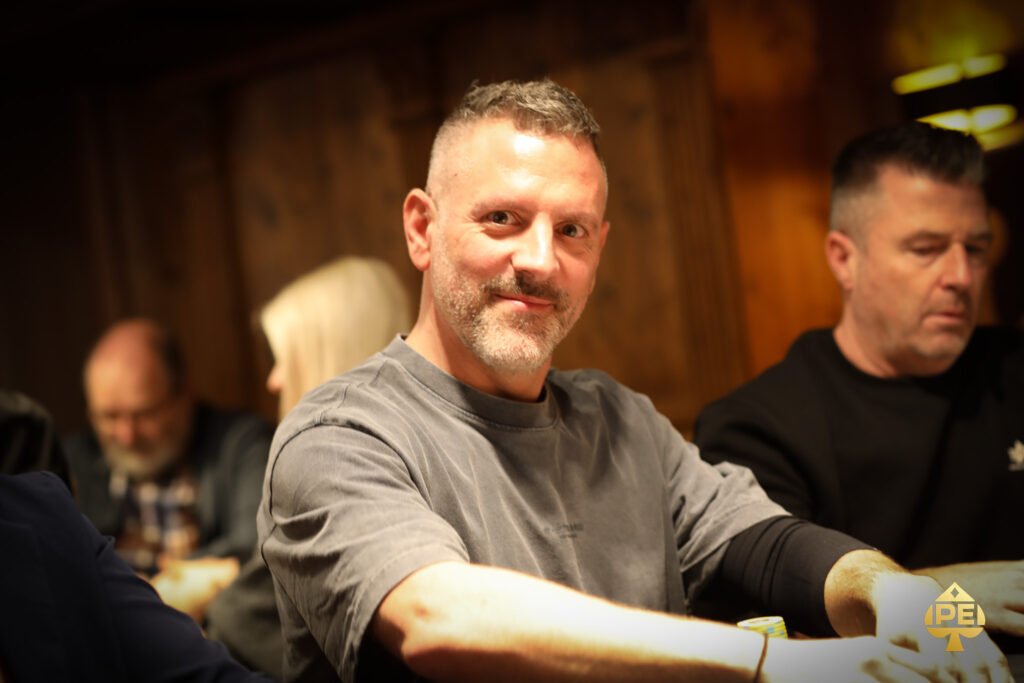 Ein Mann in einem schwarzen Anzug und einer schwarzen Sonnenbrille spielt Poker an einem Tisch. Er hält Karten in der Hand und schaut konzentriert auf den Tisch. Um ihn herum sitzen