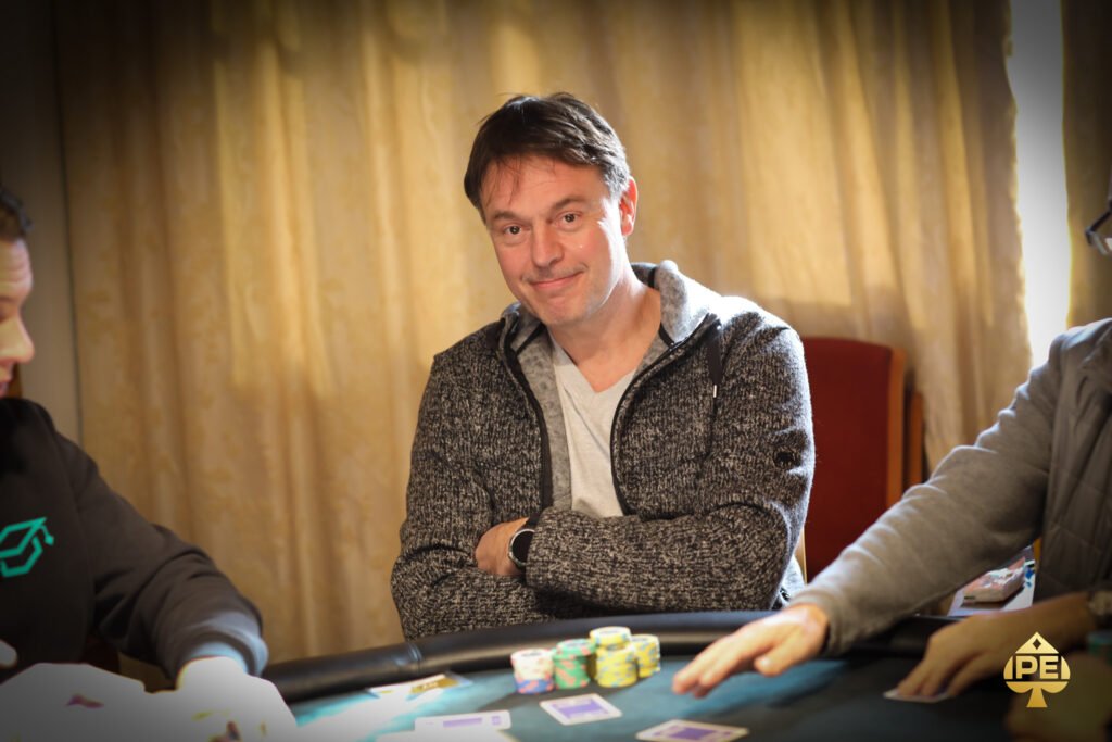 Ein Mann in einem dunkelblauen Anzug und einer Krawatte steht vor einem Pokertisch, auf dem Karten und Pokerchips liegen. Er hält zwei Karten in der Hand und schaut konzentriert auf