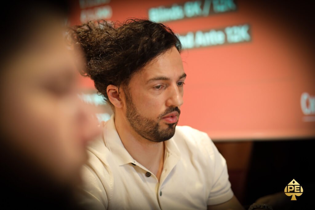 Ein Mann in dunkler Kleidung sitzt an einem Pokertisch und hält Karten in der Hand, während er konzentriert auf seine Gegner schaut. Hinter ihm sind Casino-Chips und Getränke zu