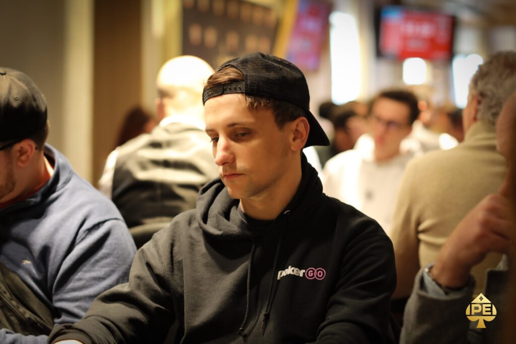 Ein professioneller Pokerspieler mit Sonnenbrille und Kappe sitzt an einem Tisch und hält eine Hand mit Spielkarten. Im Hintergrund sind weitere Spieler und Pokerchips zu sehen.