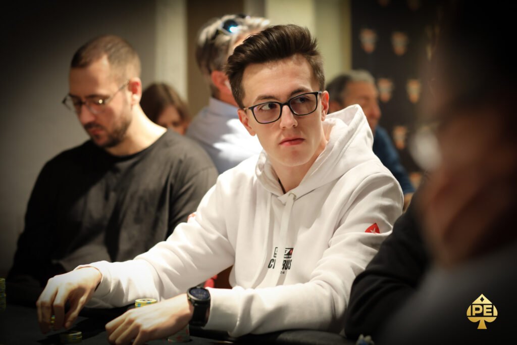Ein Mann sitzt an einem Pokertisch in einem Casino und hält zwei Karten in der Hand. Er schaut konzentriert auf den Tisch, umgeben von anderen Spielern und Pokerchips.
