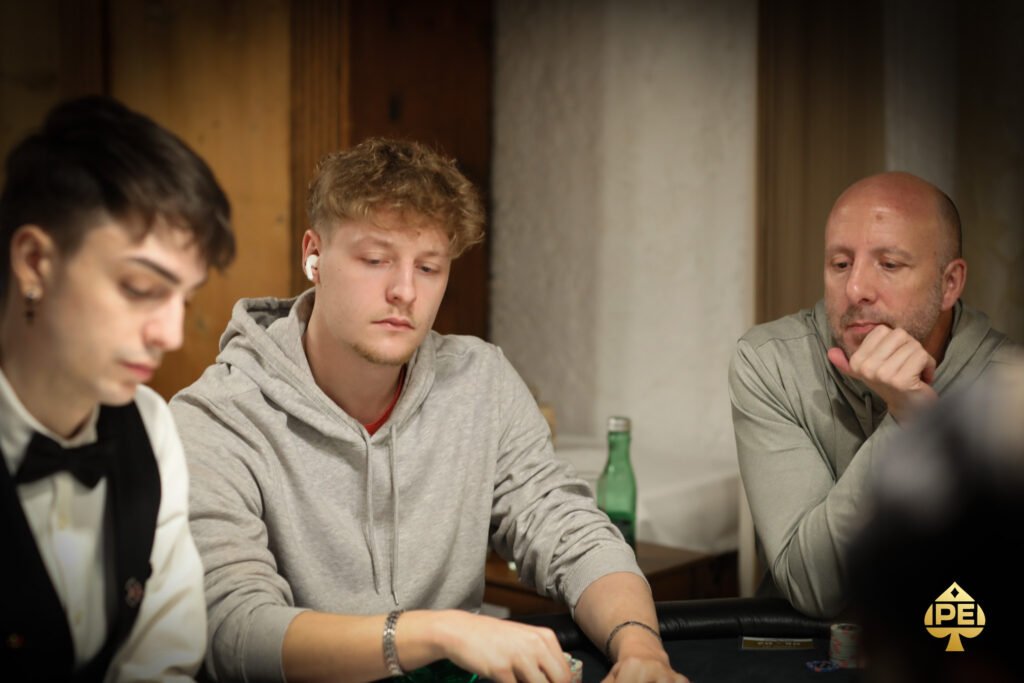 Ein Mann sitzt an einem Pokertisch und hält in seiner Hand eine Reihe von Spielkarten. Er hat einen konzentrierten Ausdruck und ist von anderen Personen umgeben, die ebenfalls Karten halten. Die Szene