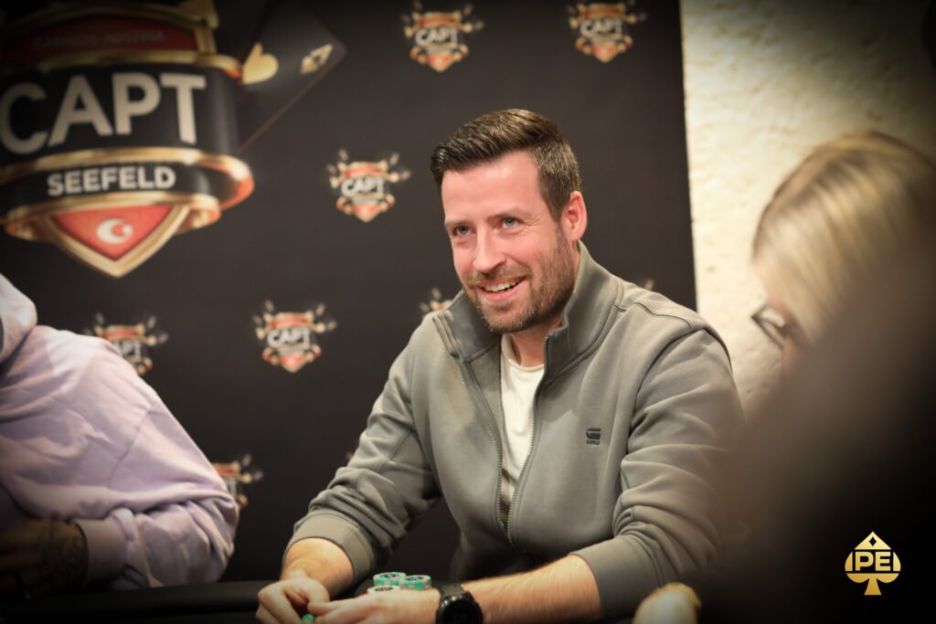 Ein Mann mit einem dunkelblauen Anzug und einer schwarzen Krawatte steht vor einem Pokertisch und hält zwei Spielkarten in der Hand. Im Hintergrund sind Pokerchips und andere Spieler zu sehen.