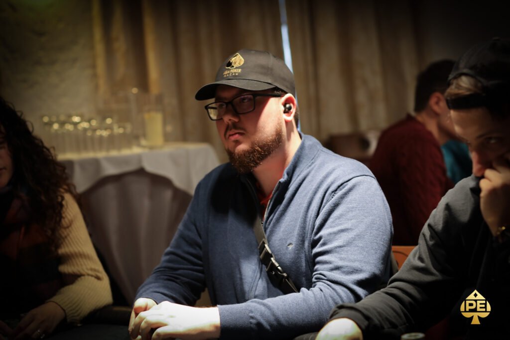 Ein Mann sitzt an einem Pokertisch mit Chips und Karten vor sich. Er hat eine ernste Miene und trägt ein dunkles Sakko. Andere Spieler sind im Hintergrund zu sehen.