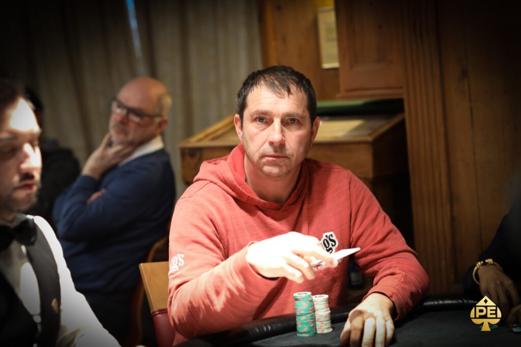 Ein Mann sitzt an einem Pokertisch, der mit Chips und Karten gefüllt ist. Er betrachtet nachdenklich seine Karten, während andere Spieler im Hintergrund spielen. Die Atmosphäre ist konzent