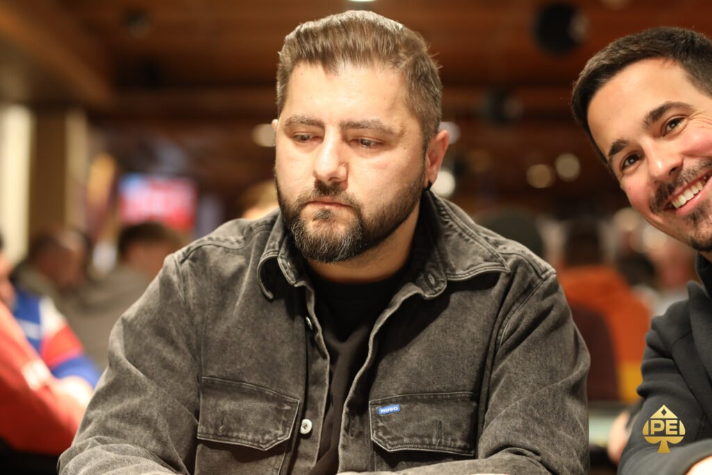 Ein Mann in einem eleganten Anzug und einer Fliege sitzt an einem Pokertisch und hält zwei Spielkarten in der Hand, während er konzentriert auf seine Gegner blickt.