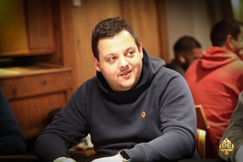 Ein Mann sitzt an einem Pokertisch und betrachtet nachdenklich seine Karten, während er von anderen Spielern umgeben ist.
